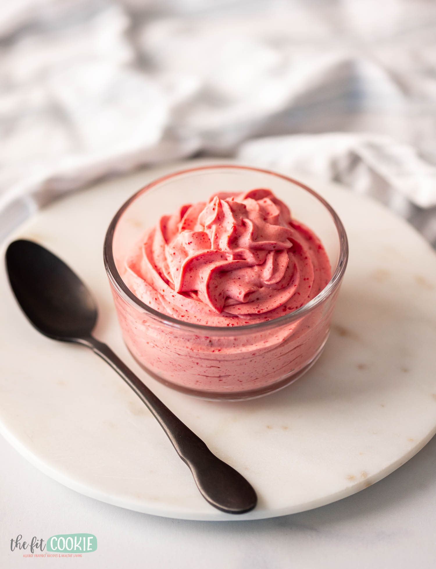 A bowl of pink strawberry mousse on a marble coaster with a spoon on the side.