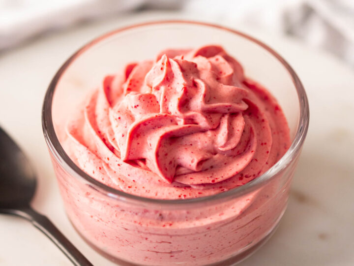 A bowl of pink whipped dessert garnished with specks, accompanied by a spoon on a white surface.
