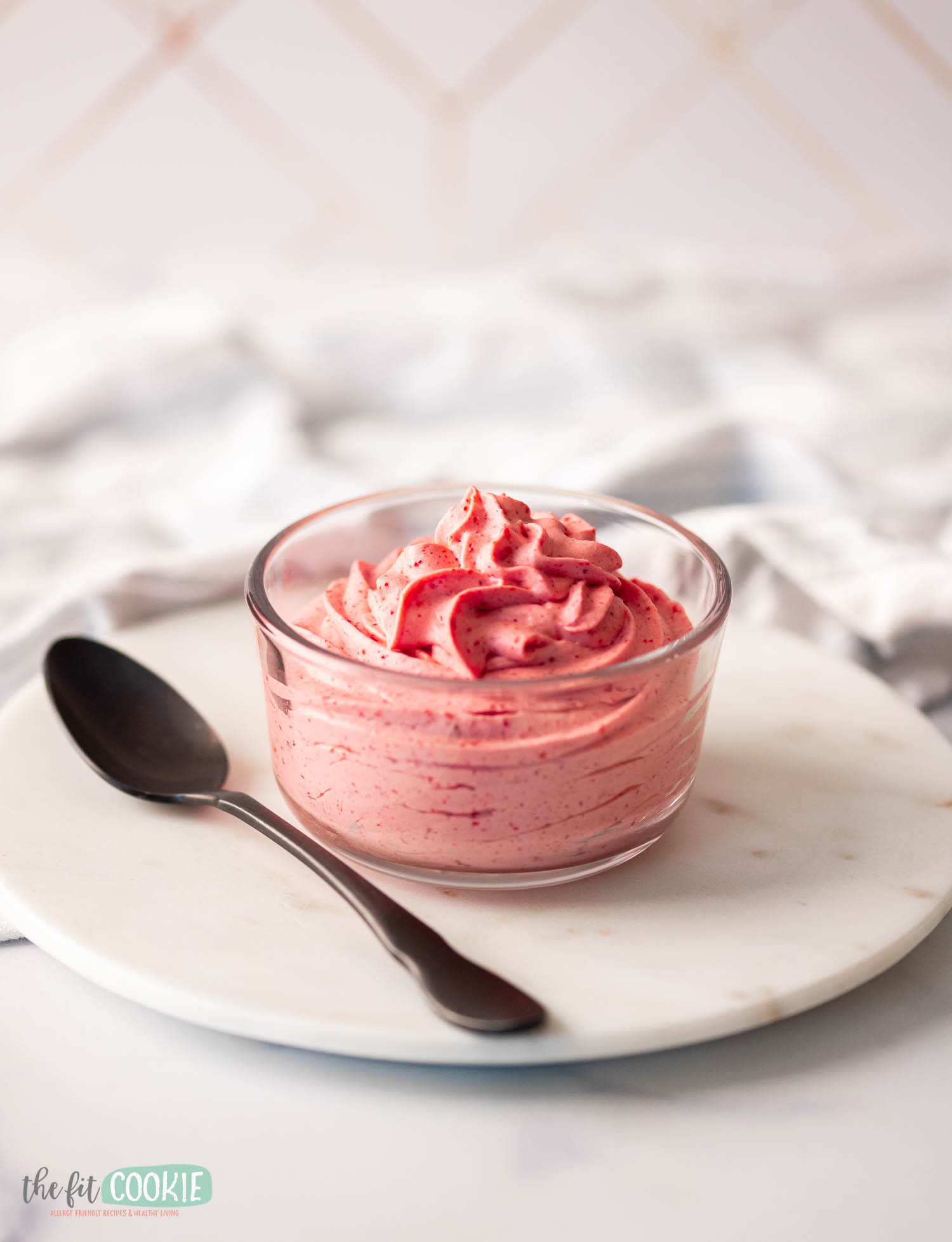 A bowl of pink strawberry mousse with a spoon on a white marble surface.