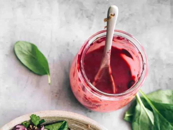 A jar of blueberry dressing with a spoon, surrounded by fresh green basil leaves.