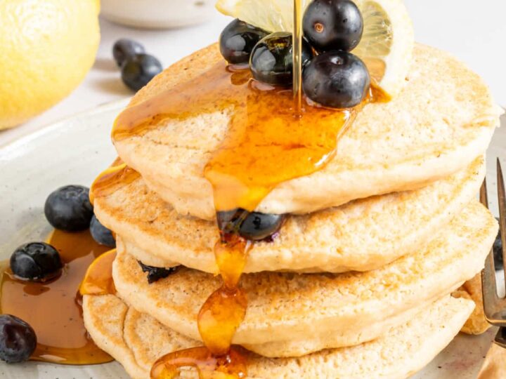 A stack of pancakes topped with blueberries and syrup being poured over, with lemon slices on the side, perfect for trying out new blueberry recipes.