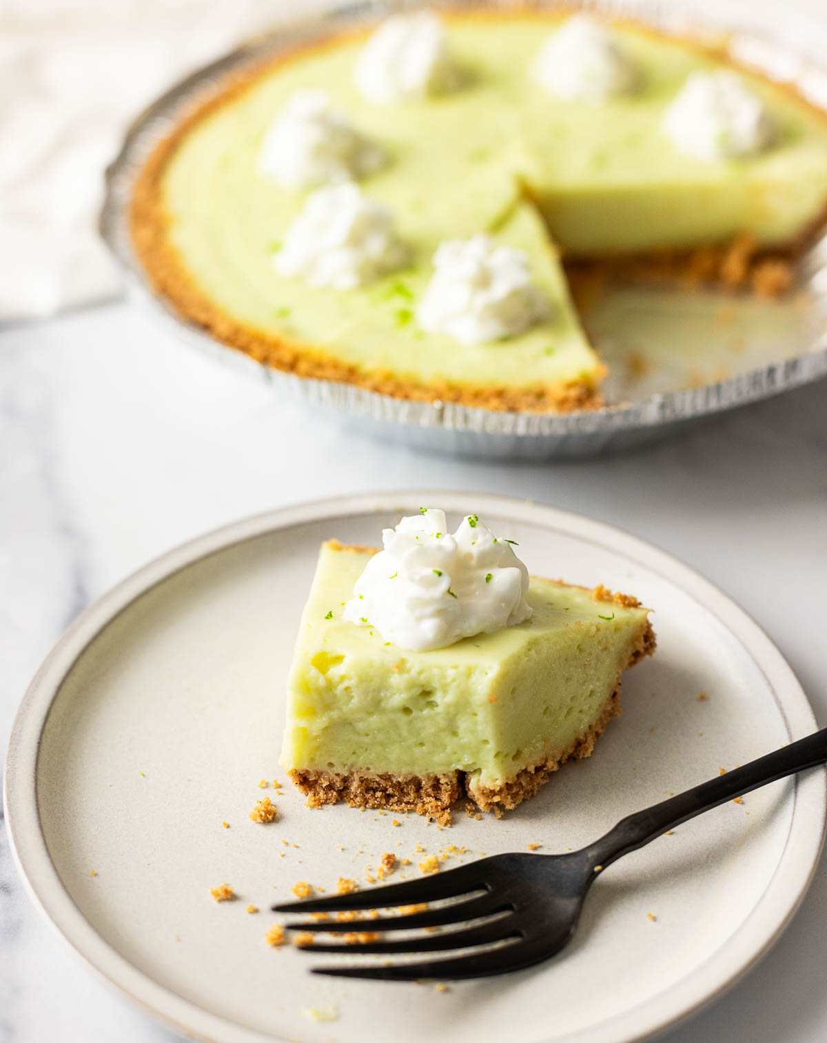 A slice of key lime pie with whipped cream on a plate, with the rest of the key lime pie in the background.
