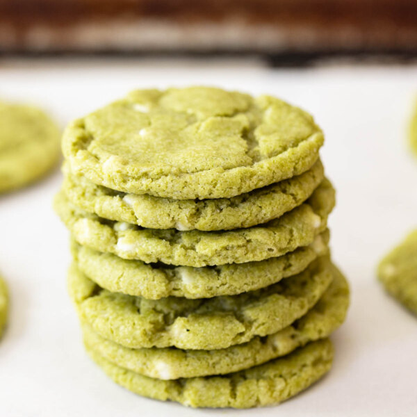 Stack of gluten free matcha cookies on a white surface with more cookies and a baking tray in the background.