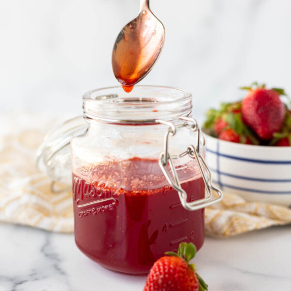 A jar of homemade strawberry simple syrup with a spoon dripping syrup into it.