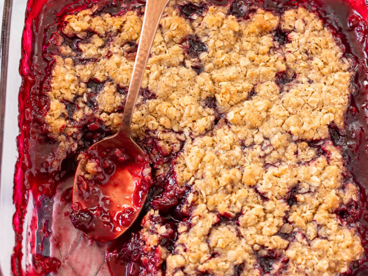 A gluten-free cherry crisp in a glass baking dish with a metal spoon. The crumble topping is golden brown, and the cherry filling is visible, surrounded by a rich red sauce.