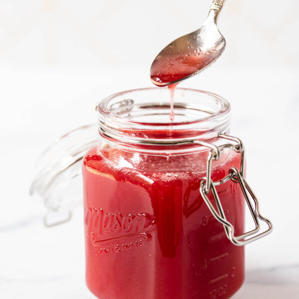 A glass Mason jar filled with rhubarb simple syrup, a spoon above the jar drips some of the syrup.