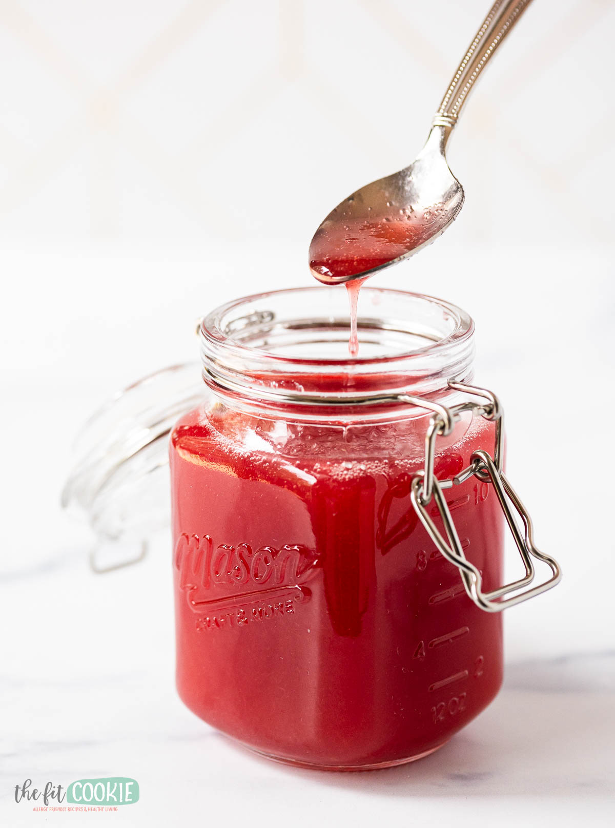 A glass Mason jar filled with rhubarb simple syrup, a spoon above the jar drips some of the syrup.