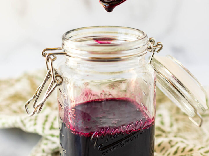 A jar of dark purple blueberry simple syrup is open with a spoon dripping some of the syrup above it. A green and white cloth is partially visible in the background.