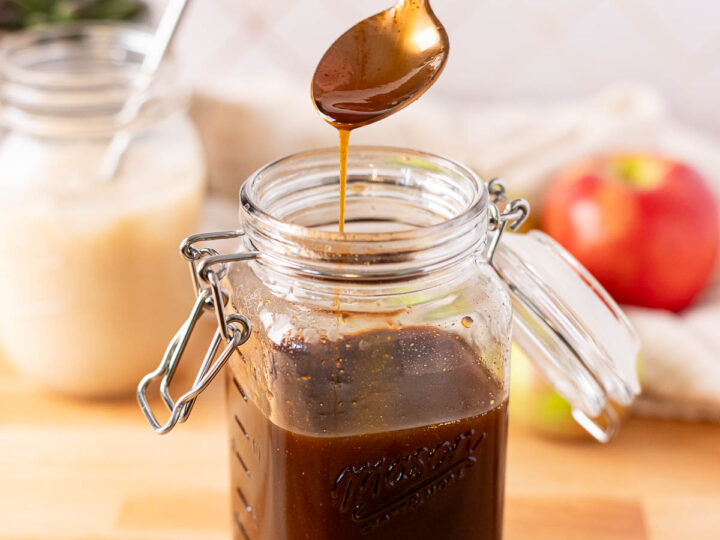 A spoon drizzles dark apple chai syrup from a mason jar on a wooden surface, with an apple and a drink in the background.