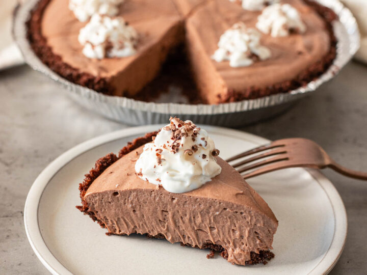 A slice of chocolate cream pie with whipped cream on a plate, featuring the whole pie in the background, offers a dairy-free delight that everyone can enjoy.