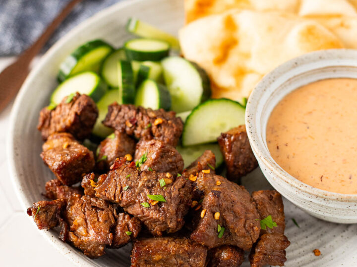 A plate with air fryer steak bites, sliced cucumber, flatbread, and a bowl of dipping sauce sits on the table alongside a fork and napkin.