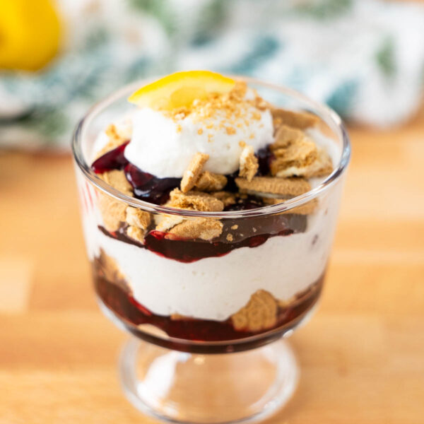 A blackberry lemon trifle dessert in a glass cup with whipped cream, crumbled cookies, berry sauce, and a lemon slice on top, placed on a wooden surface.