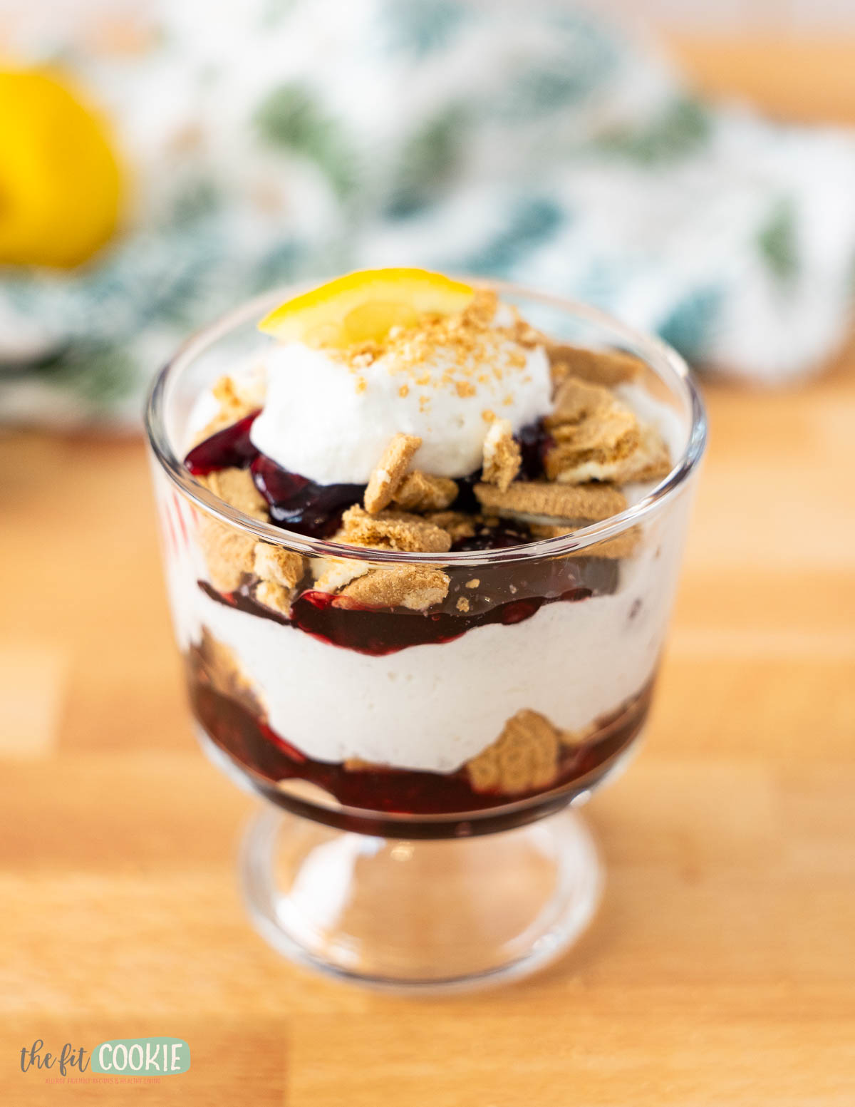 A blackberry lemon trifle dessert in a glass cup with whipped cream, crumbled cookies, berry sauce, and a lemon slice on top, placed on a wooden surface.