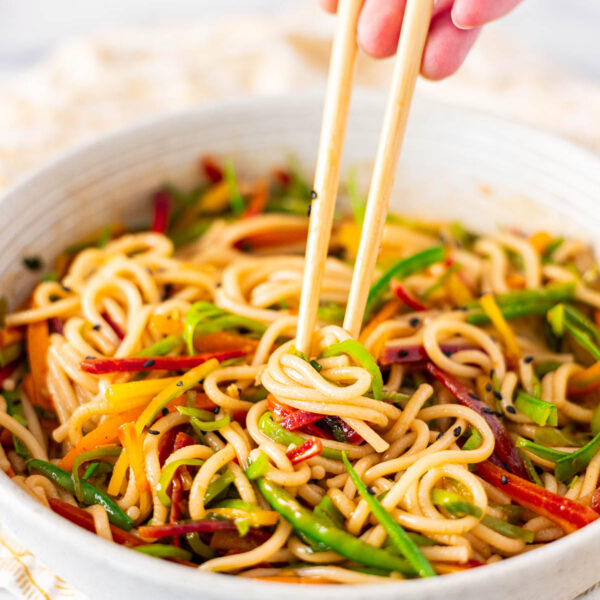 A hand uses chopsticks to pick up udon noodle salad mixed with sliced vegetables in a white bowl.