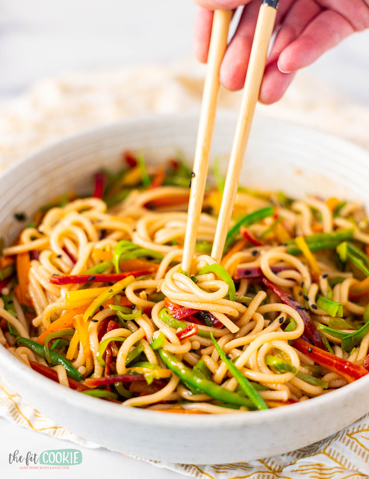 A hand uses chopsticks to pick up udon noodle salad mixed with sliced vegetables in a white bowl.