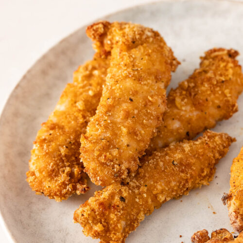 A plate with several pieces of golden-brown gluten free air fryer chicken tenders on a light-colored surface.