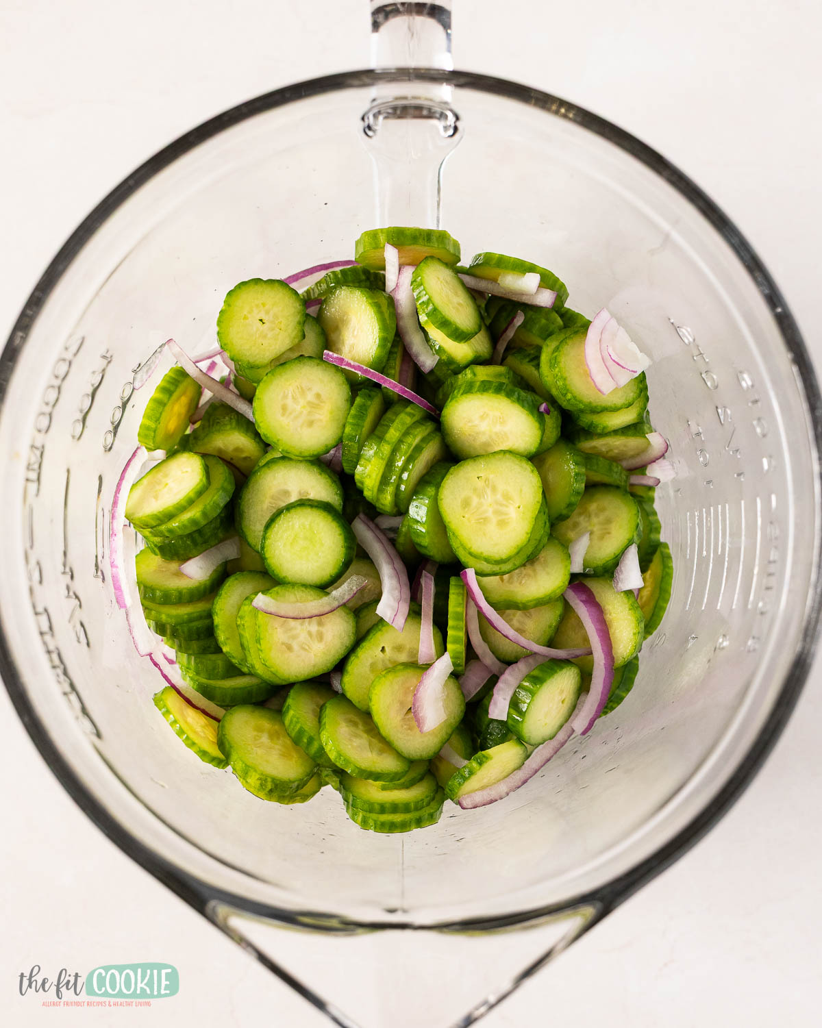 A glass mixing bowl containing cucumber salad with sliced cucumbers and thinly sliced red onions on a white surface.