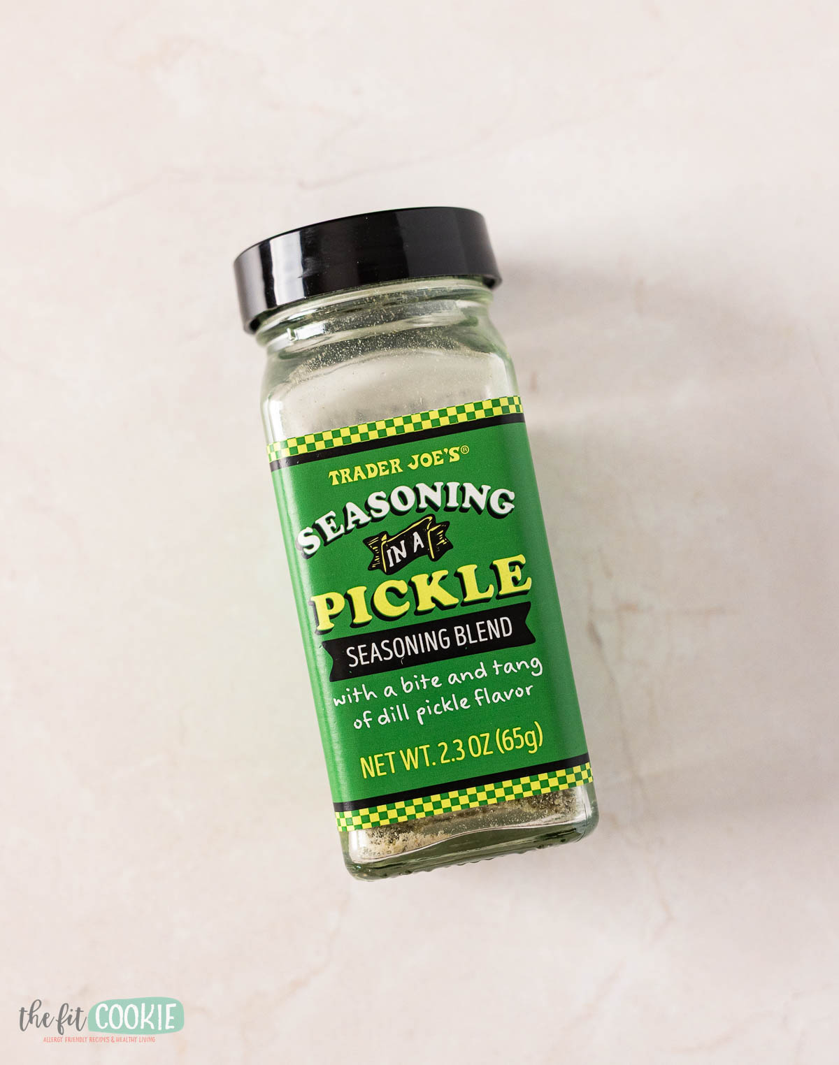 A glass jar of Trader Joe's Seasoning In A Pickle seasoning blend with a green label sits on a light-colored surface—perfect for adding tangy flavor to your next cucumber salad.