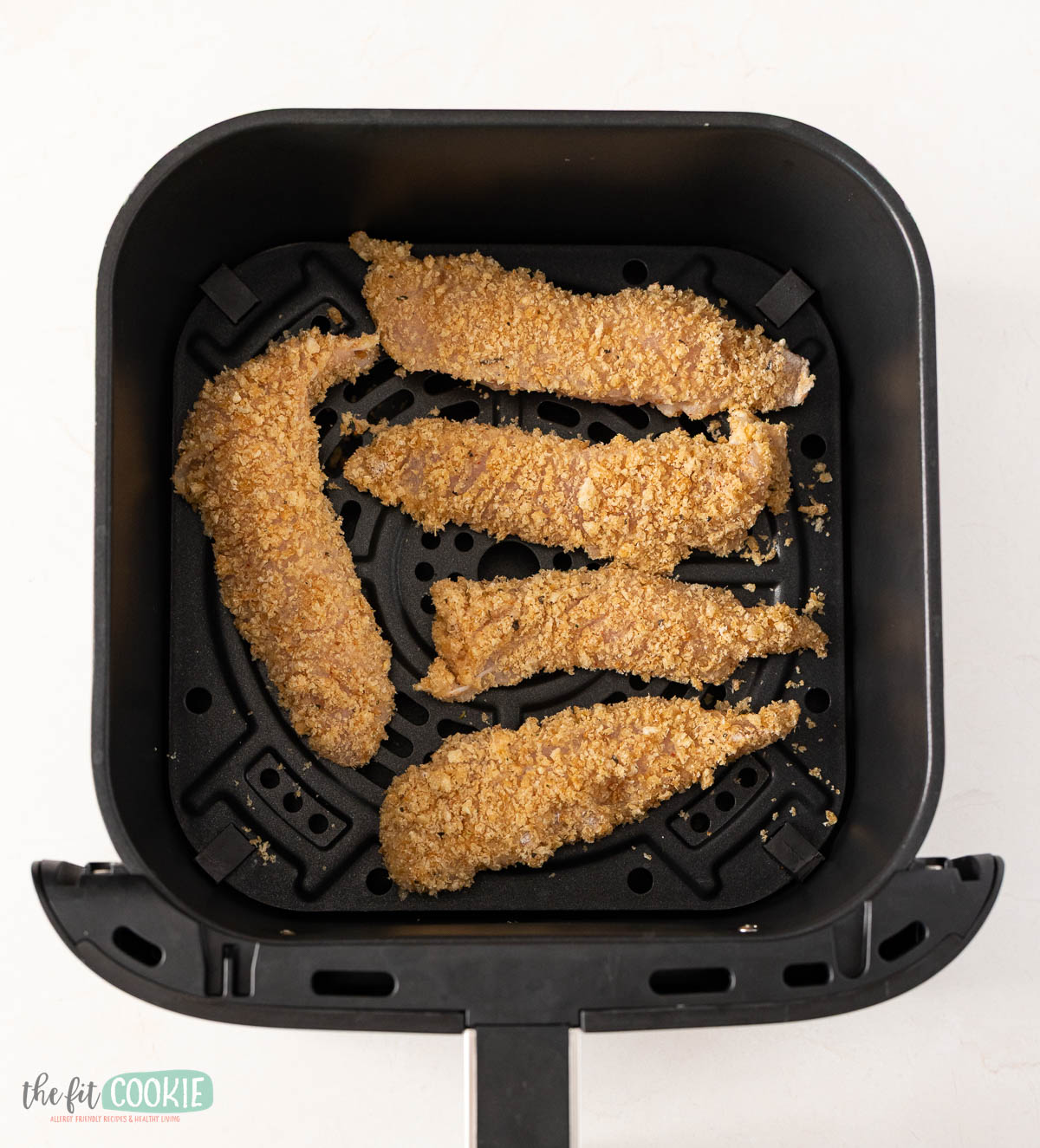 Five gluten free air fryer chicken tenders are arranged in the basket of an air fryer, ready to be cooked.