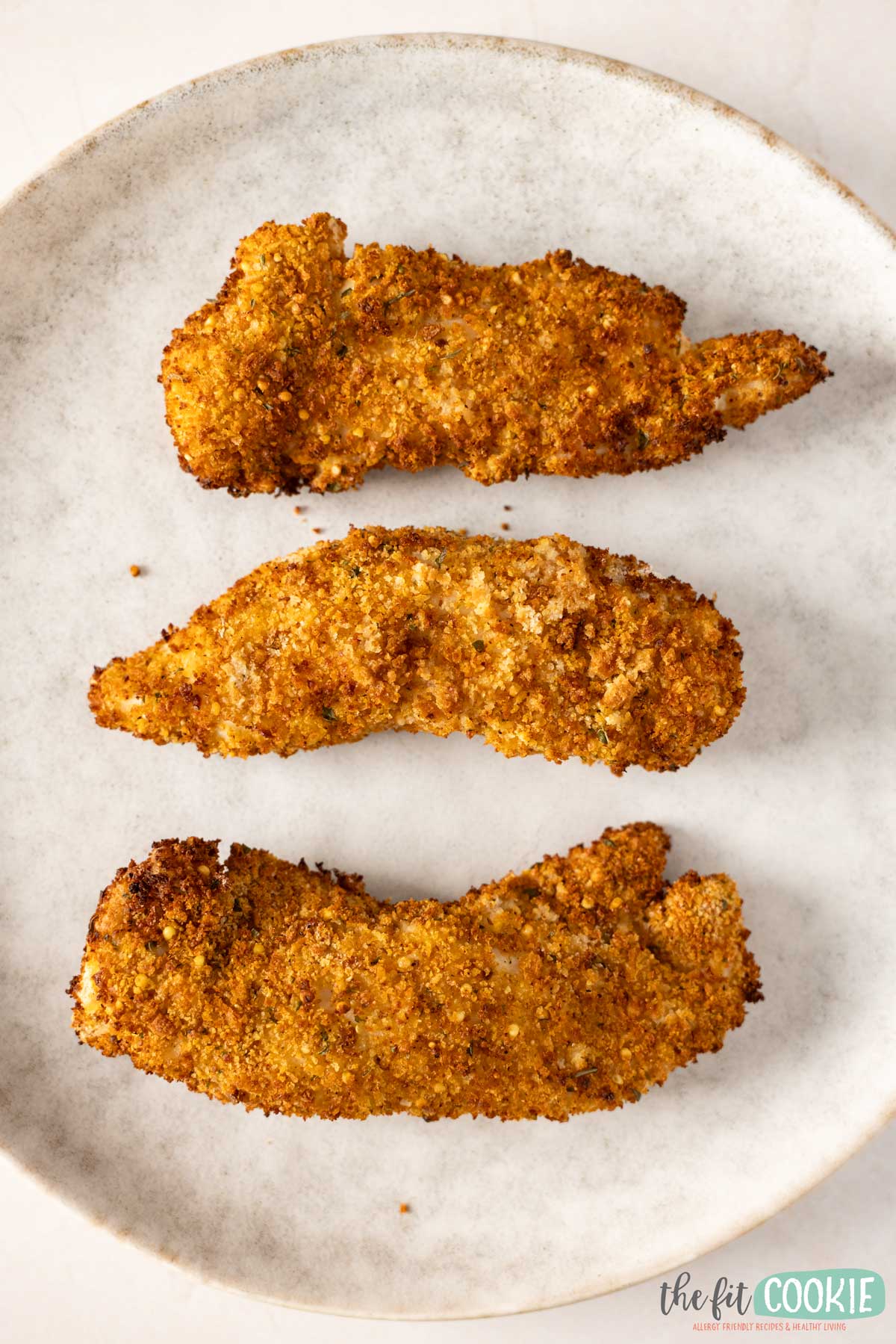 Three gluten free air fryer chicken tenders coated with homemade bread crumbs arranged in a row on a light-colored plate.