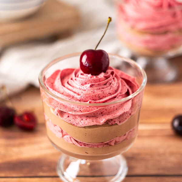A glass dessert cup filled with layers of chocolate cherry mousse and pink cherry mousse, topped with a whole cherry.