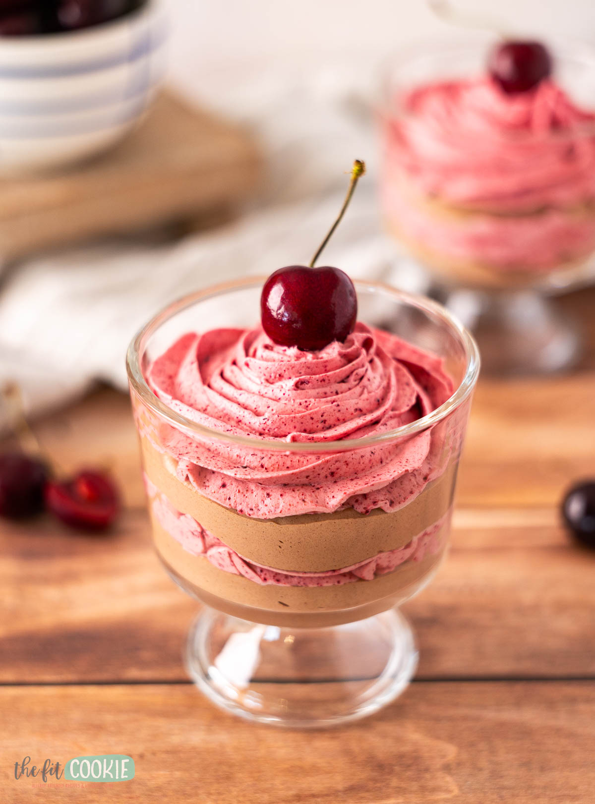 A glass dessert cup filled with layers of chocolate cherry mousse and pink cherry mousse, topped with a whole cherry.