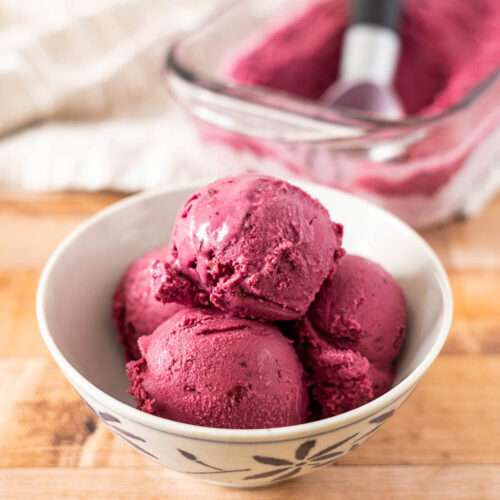 A white bowl with four scoops of dark purple, dairy free huckleberry ice cream sits on a wooden surface, with a glass dish of sorbet and an ice cream scoop in the background.
