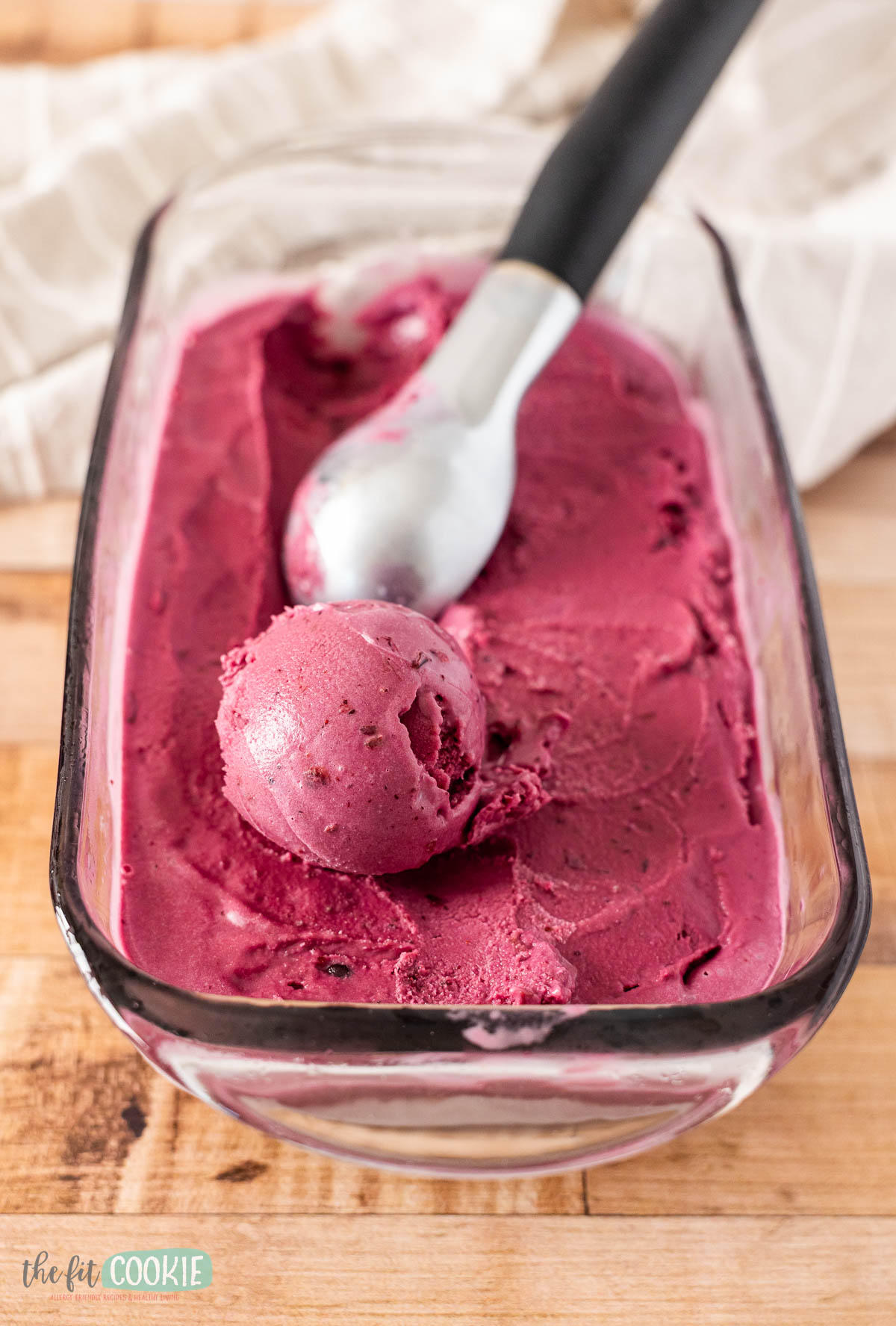 A glass container of dark pink dairy free huckleberry ice cream with a metal scoop and one round scoop of ice cream on top.