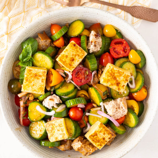 A vibrant panzanella salad featuring grilled tofu cubes, sliced cucumbers, cherry tomatoes, red onions, bread chunks, and basil is served in a bowl on a patterned napkin with a fork.