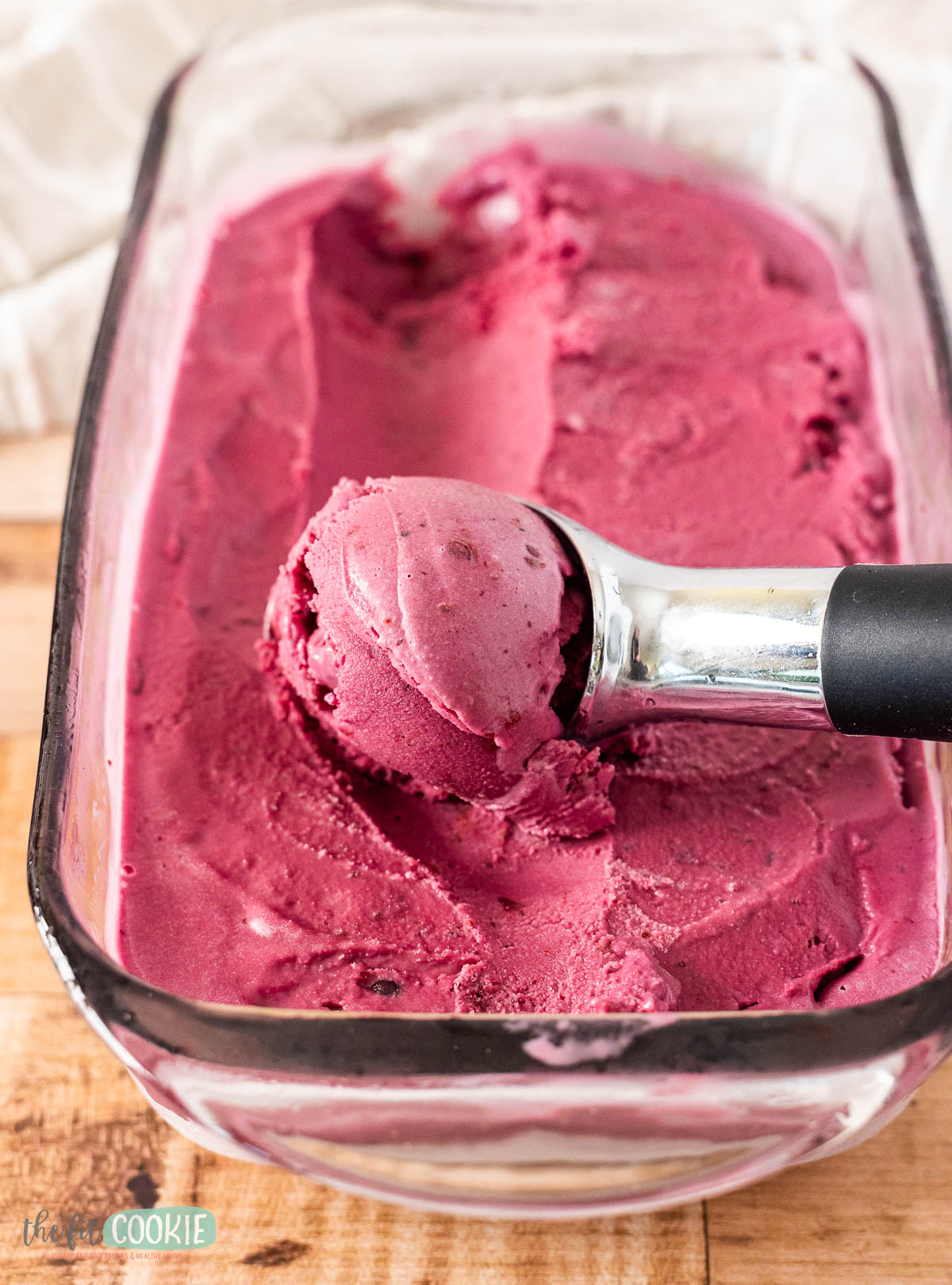 A metal ice cream scoop in a glass container holds dairy free huckleberry ice cream, with some of the rich, dark purple treat already scooped out.