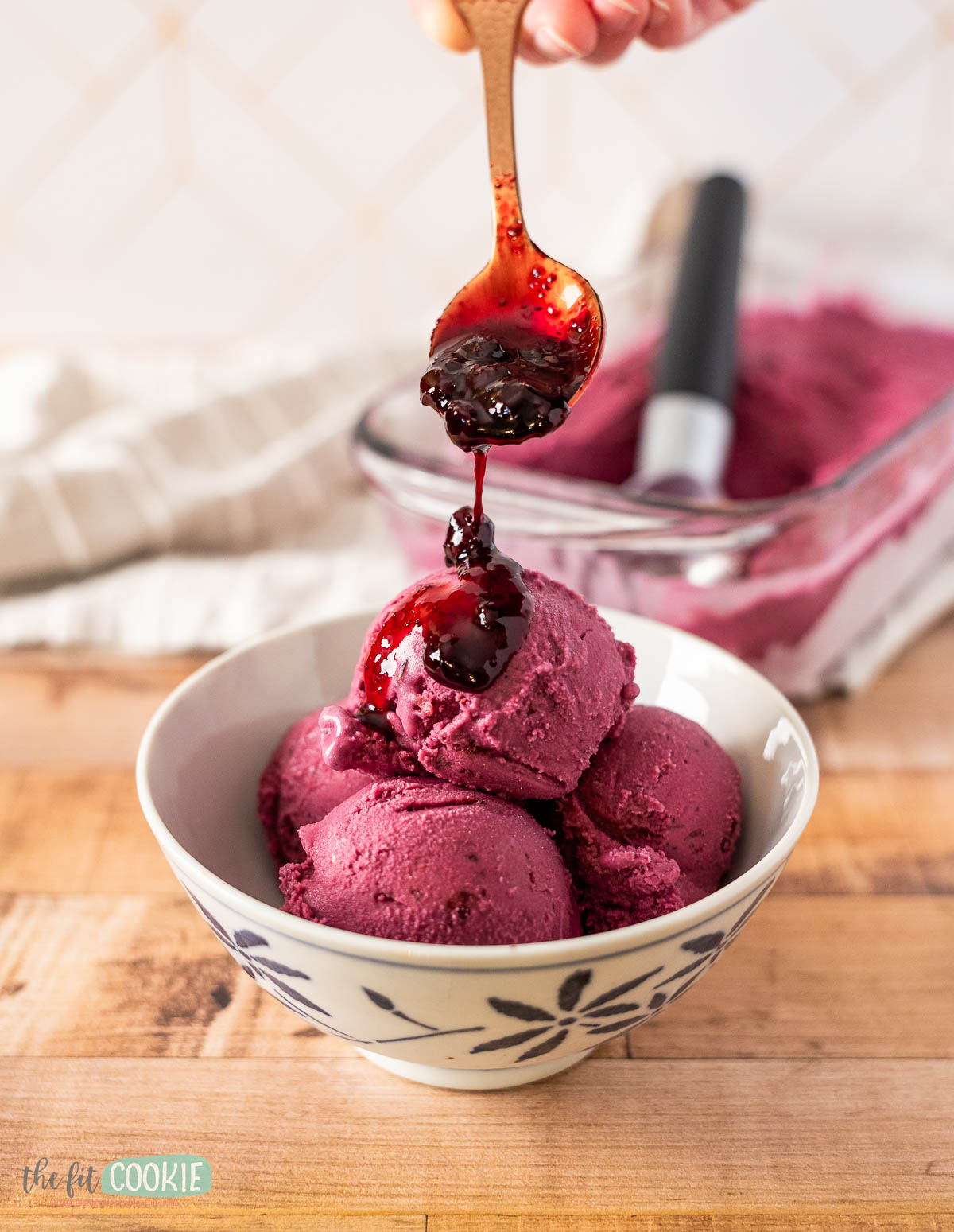 A hand holds a spoon, drizzling rich huckleberry sauce over scoops of dairy free huckleberry ice cream.