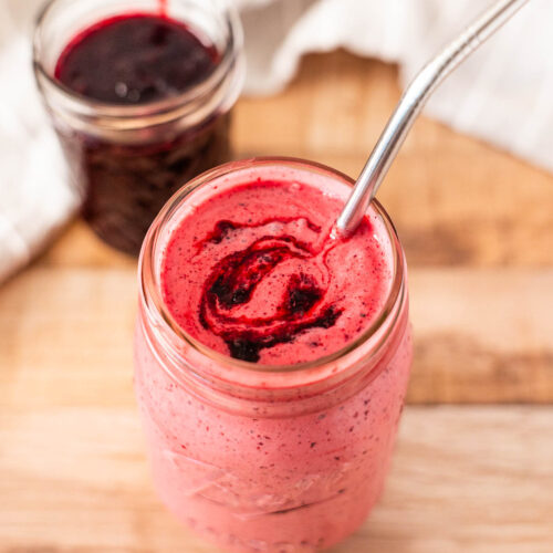 A dairy free mason jar milkshake, filled with a pink smoothie swirled with dark huckleberry sauce, sits on a wooden surface next to a small jar of berry jam.