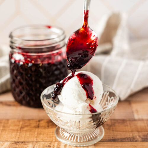 A spoon pours huckleberry sauce over a scoop of vanilla ice cream in a glass bowl, with a jar of berry sauce and a cloth in the background.