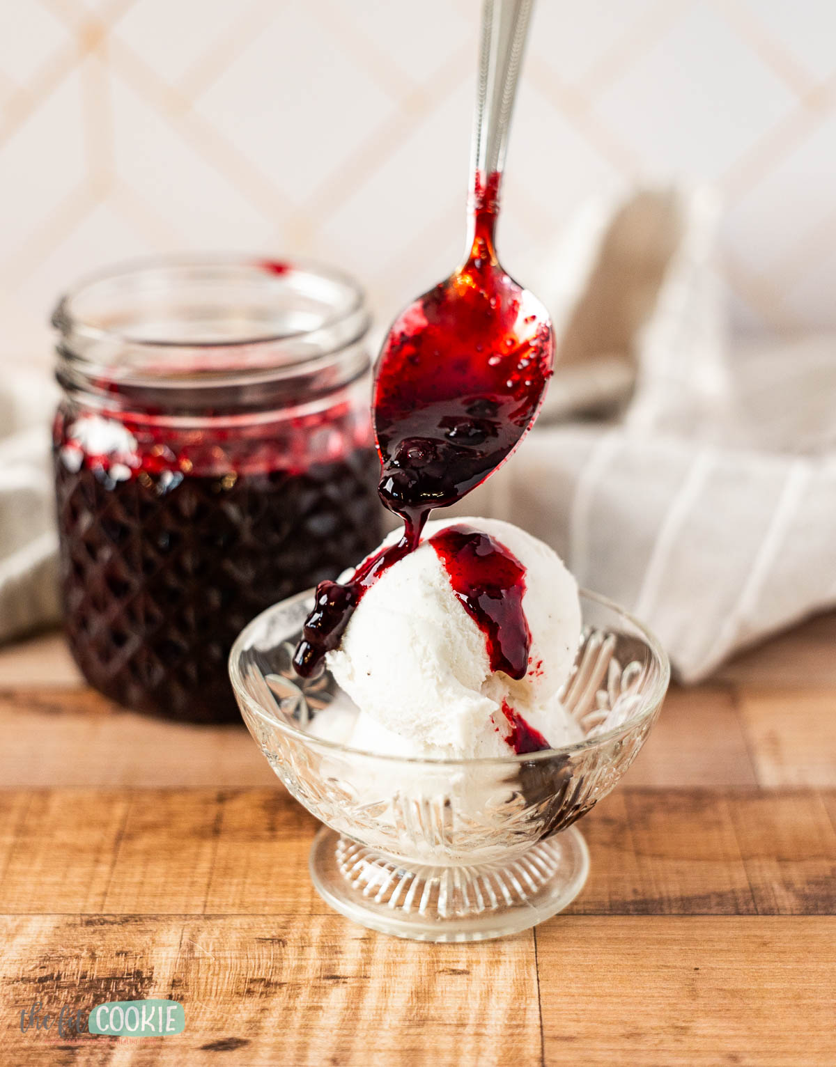 A spoon pours huckleberry sauce over a scoop of vanilla ice cream in a glass bowl, with a jar of berry sauce and a cloth in the background.
