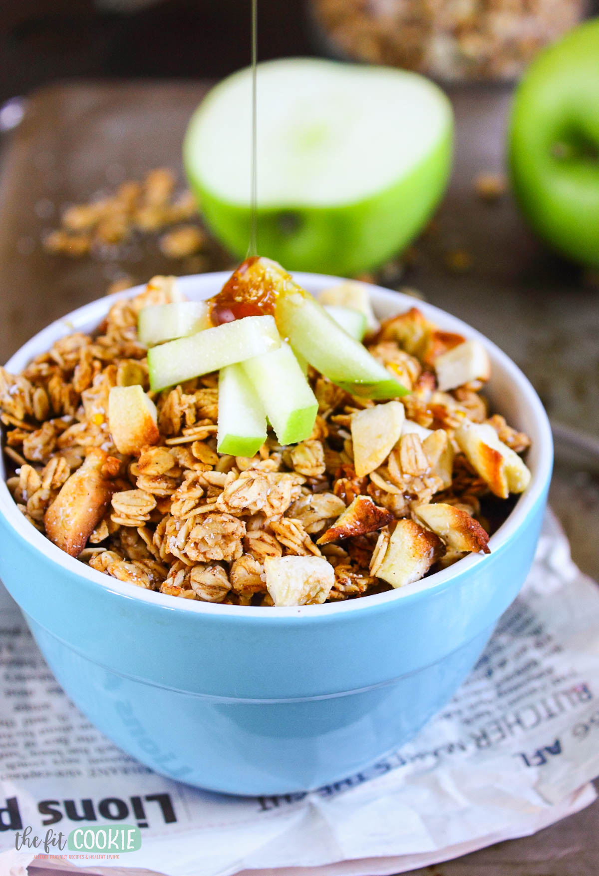 A bowl of apple cinnamon granola topped with fresh apple slices, with syrup being drizzled on top; a green apple and extra granola are visible in the background.