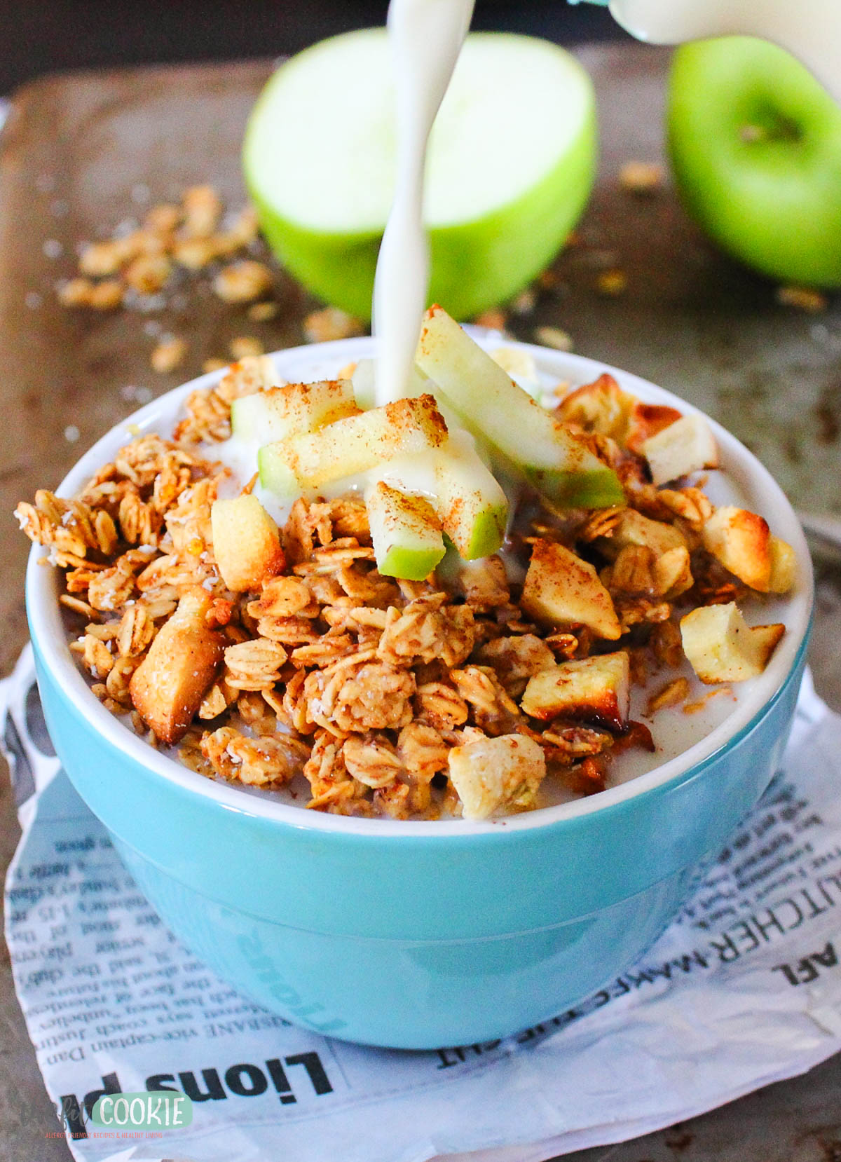A bowl of apple cinnamon granola with chopped green apples is topped with milk being poured, set on newspaper with a green apple in the background.