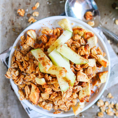 A bowl of apple cinnamon granola topped with fresh apple slices and a sprinkle of cinnamon, with a spoon and scattered granola pieces nearby.
