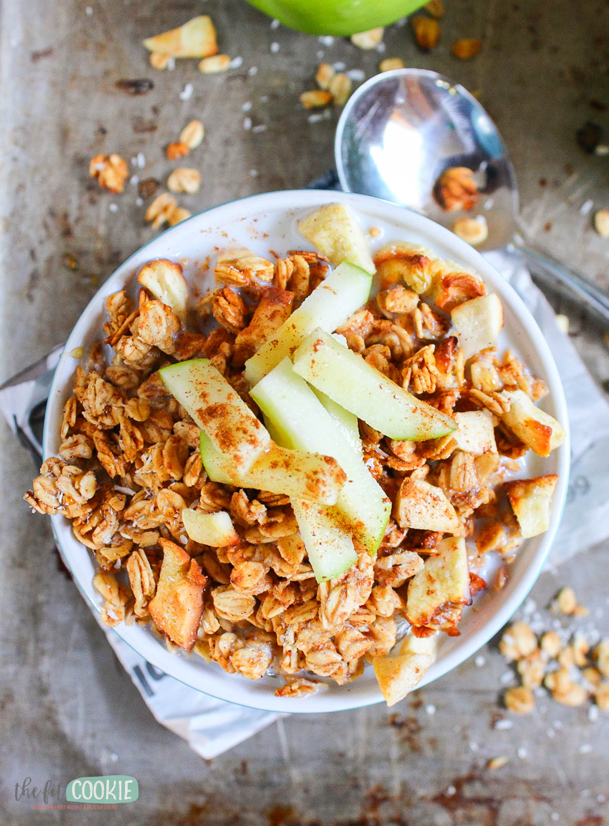 A bowl of apple cinnamon granola topped with fresh apple slices and a sprinkle of cinnamon, with a spoon and scattered granola pieces nearby.