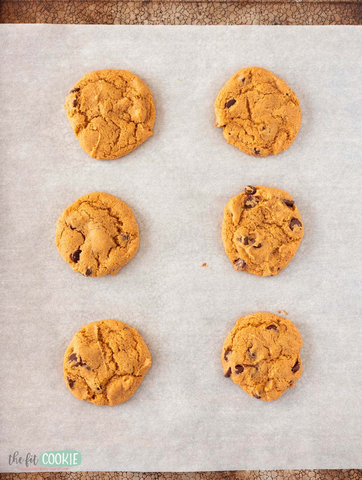 Six pumpkin chocolate chip cookies are arranged in two columns on sheet pan covered in parchment paper.