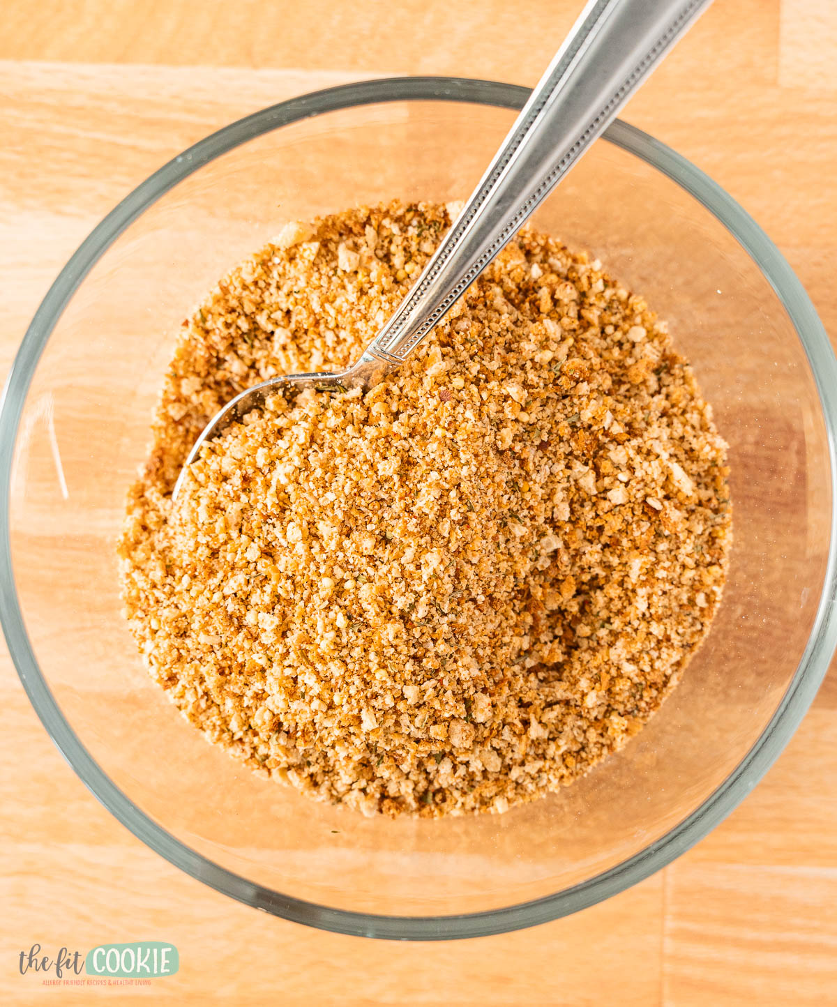 A clear glass bowl filled with gluten free seasoned bread crumbs and a metal spoon, placed on a wooden surface.