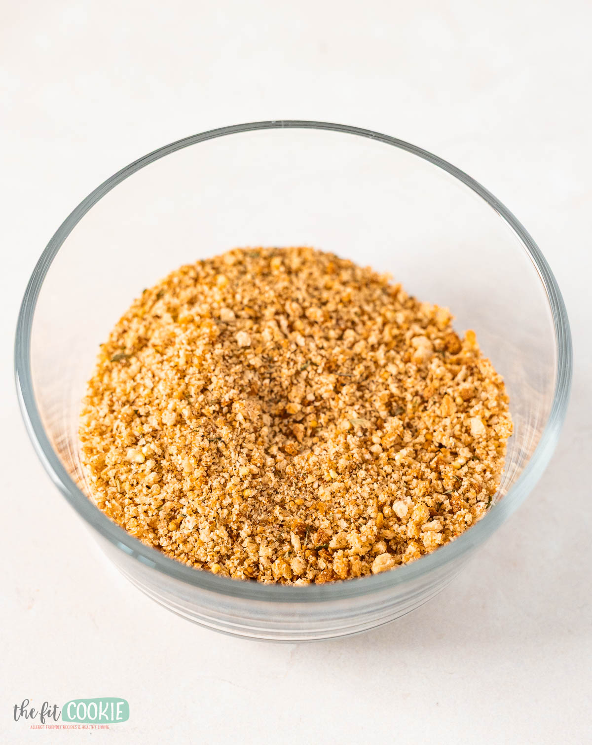 A clear glass bowl filled with finely crushed gluten free seasoned bread crumbs on a light surface.