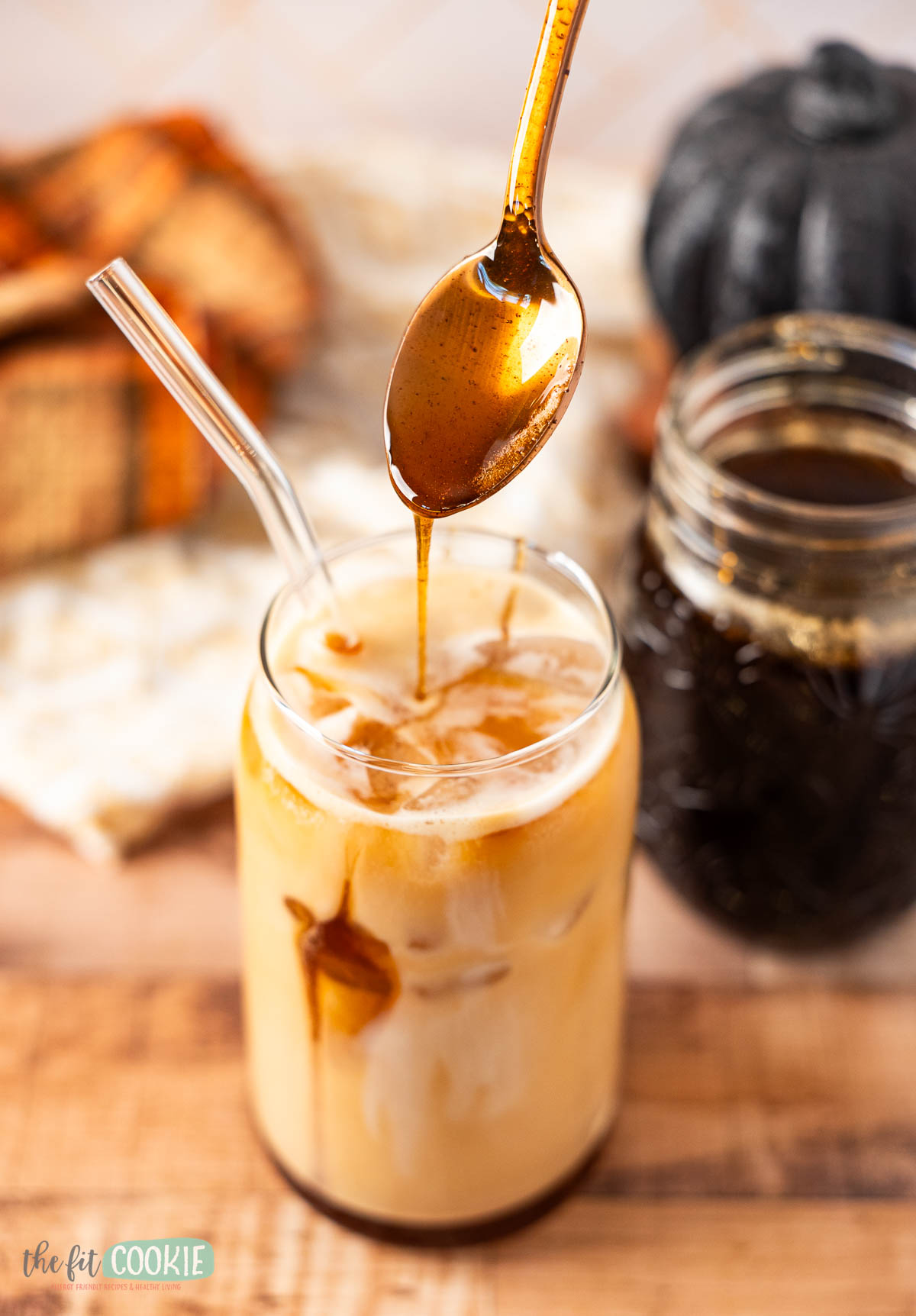 A glass of iced coffee with a glass straw is being drizzled with homemade brown sugar syrup from a spoon; a mason jar of syrup and a pumpkin are in the background.