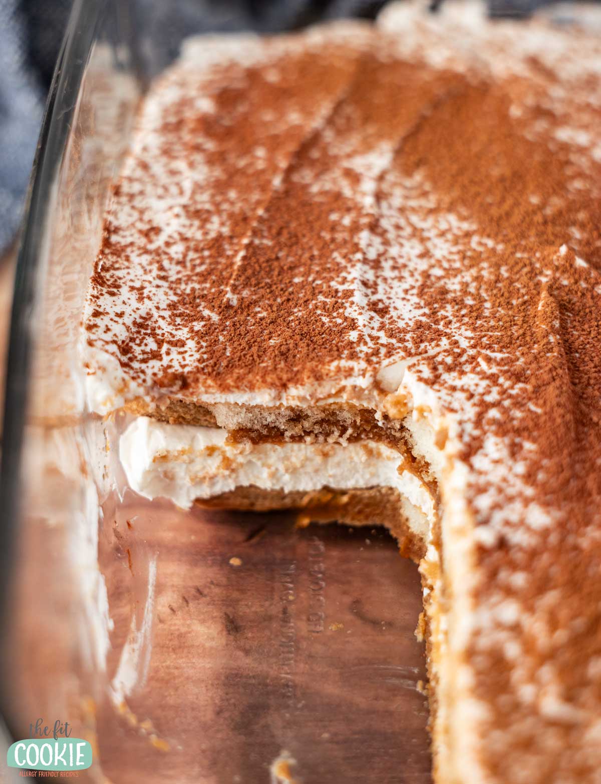 A rectangular glass dish of dairy free tiramisu with a section removed, revealing creamy layers topped with cocoa powder.