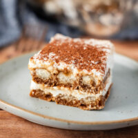 A square slice of dairy free tiramisu dessert with layers of creamy filling and coffee-soaked cake, topped with cocoa powder, served on a light-colored plate.
