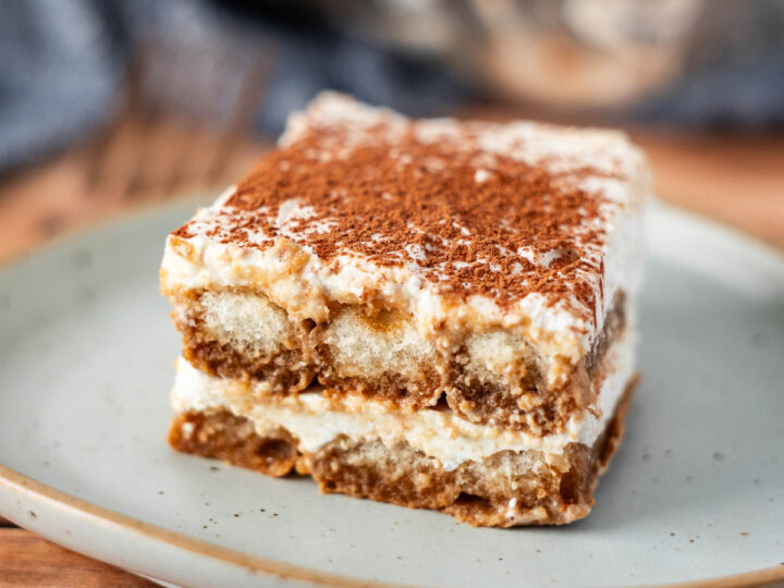 A square slice of dairy free tiramisu dessert with layers of creamy filling and coffee-soaked cake, topped with cocoa powder, served on a light-colored plate.