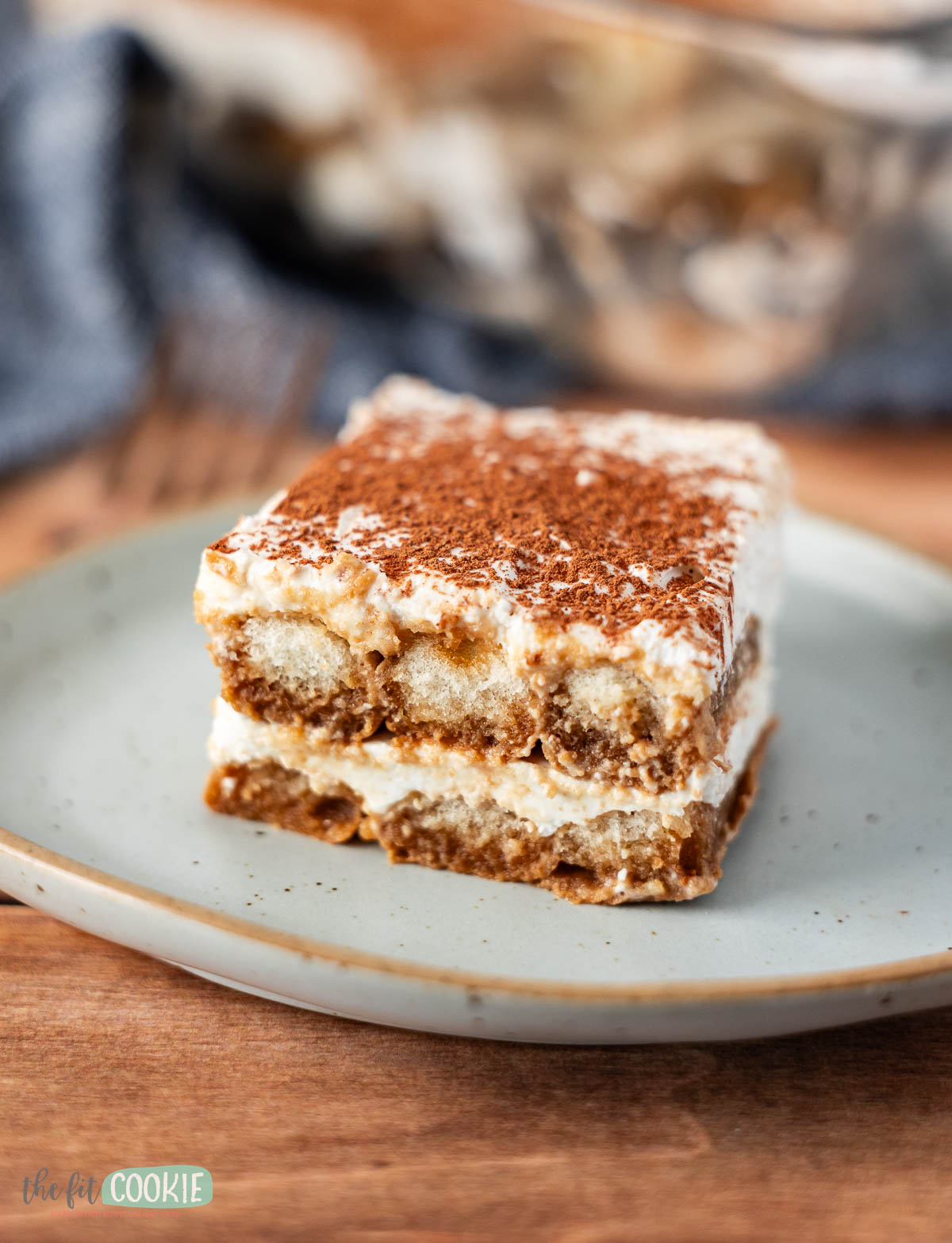 A square slice of dairy free tiramisu dessert with layers of creamy filling and coffee-soaked cake, topped with cocoa powder, served on a light-colored plate.