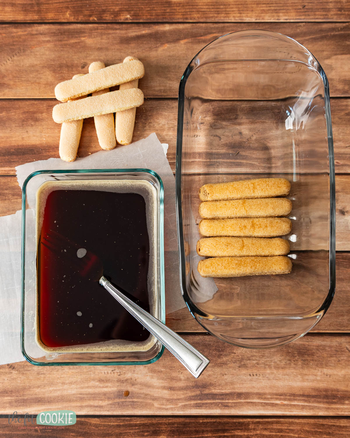 A glass dish with dairy free tiramisu, a container of coffee with a spoon, and several ladyfingers on a napkin are arranged on a wooden surface.