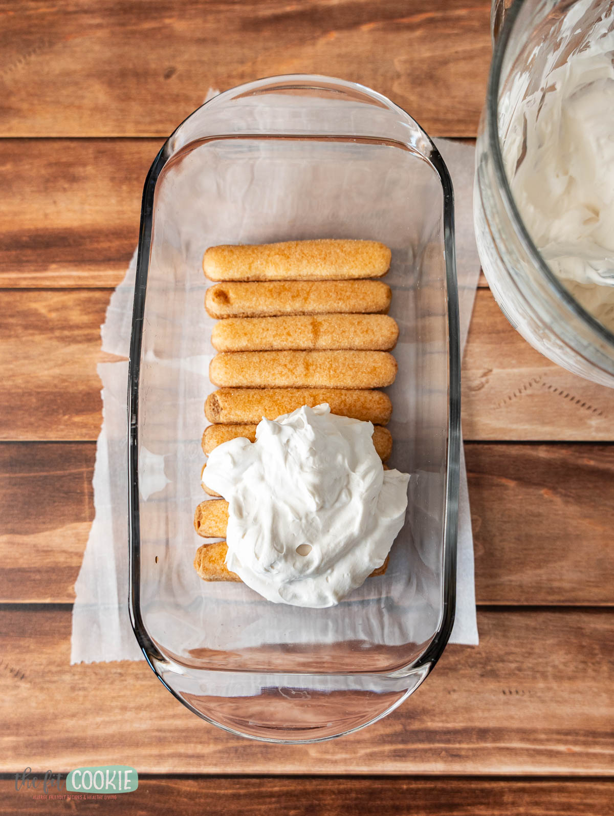 A glass dish of dairy free tiramisu, featuring a layer of ladyfinger cookies topped with a generous dollop of whipped cream, sits on a wooden surface next to a mixing bowl.
