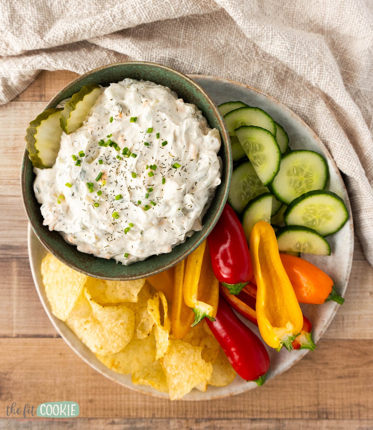 A bowl of creamy dill pickle dip topped with herbs and pickle slices, served with potato chips, sliced cucumbers, and mini bell peppers on a plate.