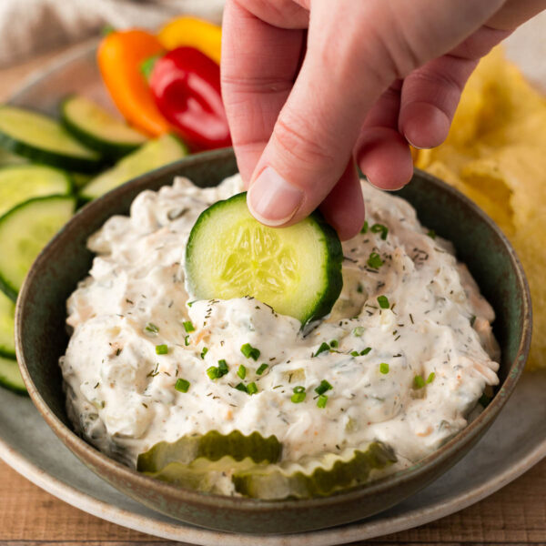 A hand dips a cucumber slice into a bowl of creamy dill pickle dip, with sliced cucumbers, mini bell peppers, and chips on the side.